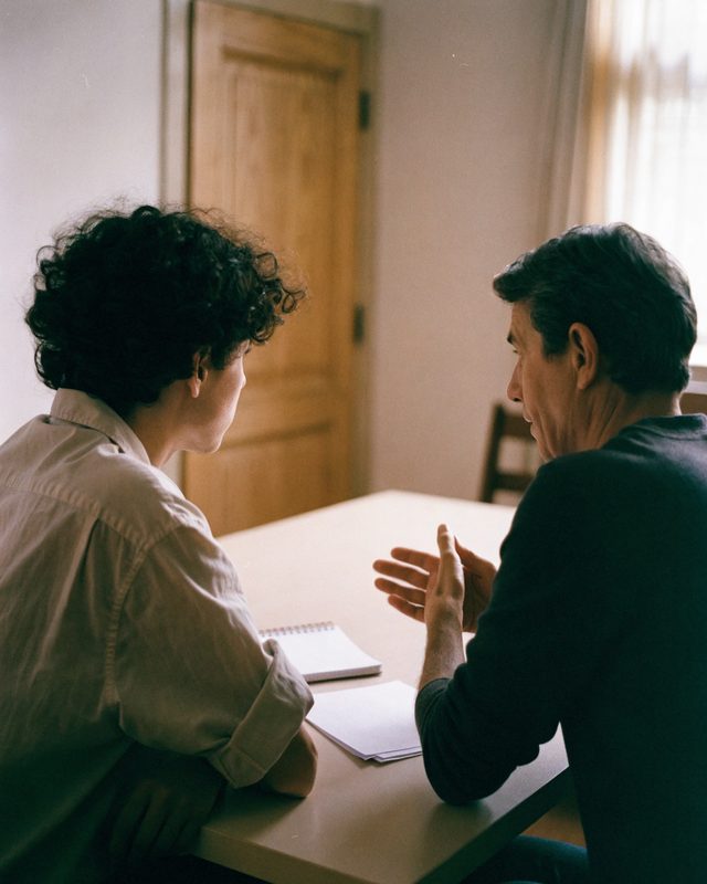 Two people at a plain table, one explaining something while the other listens.