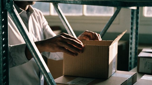 Hands packing a box on warehouse shelving in morning light.